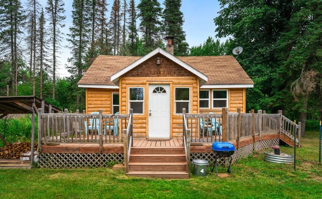 Small log cabin with wraparound deck, grill, and firewood surrounded by tall pine trees
